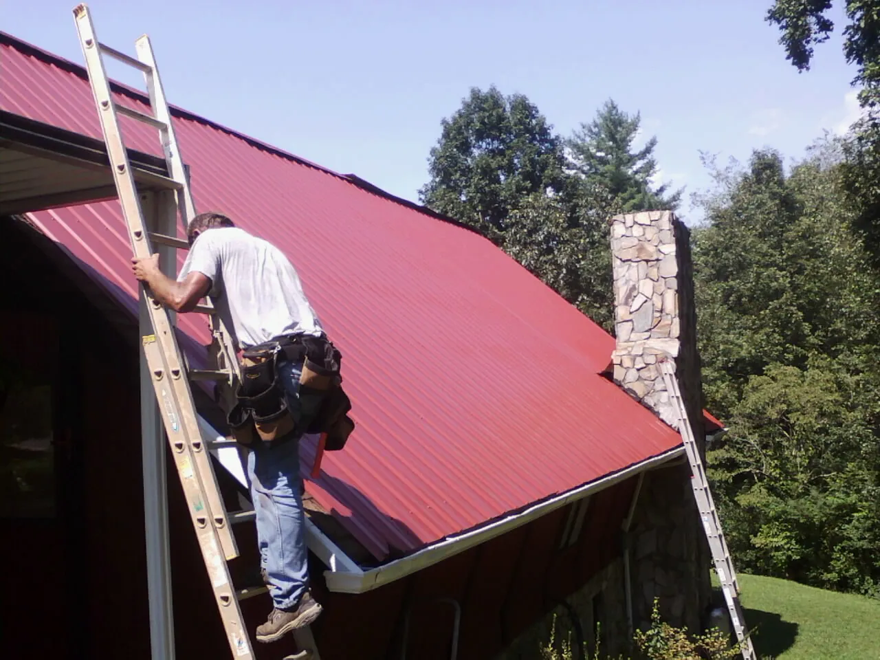 Roofer on a ladder beside a red metal roof and stone chimney — Soffit & Fascia Repair by Roofing Brothers in Avinger